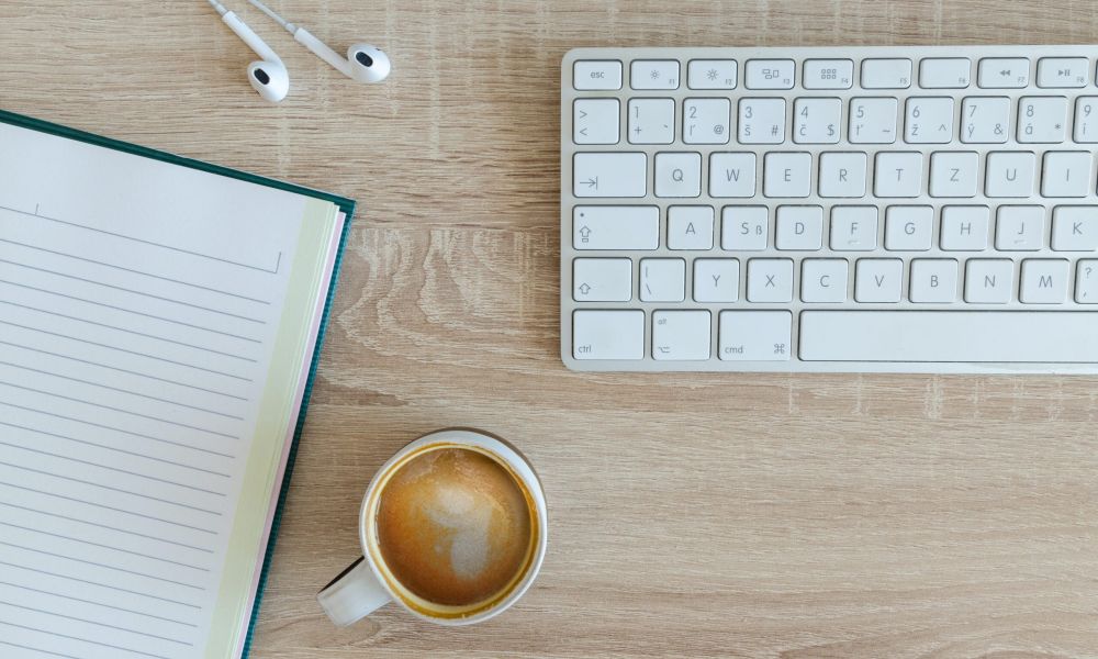 The image shows a typical workspace setup with a keyboard, a notebook, and a cup of coffee on a wooden table. It depicts a simple yet functional environment for working or studying. The open notebook suggests the possibility of taking notes or writing tasks, while the keyboard represents the use of a computer or laptop. The presence of a hot coffee adds a comforting element to the scene, providing a familiar accompaniment for many during work sessions. Overall, the image conveys a minimalist yet practical workspace setting.